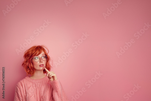 jeune femme rousse à taches de rousseurs, lunettes, qui doute et pose son index devant sa bouche pour montrer son hésitation, elle porte un pull en laine rose à torsades, sur fond rose avec copyspace