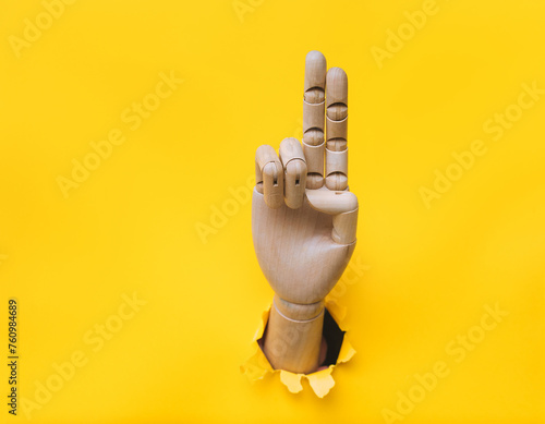A wooden hand protrudes from a torn hole in yellow paper and shows two fingers. Concept of Catholicism, savior gesture, Christianity.