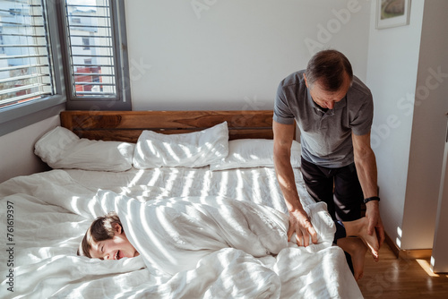 Grandpa playing with his little grandson wrapping him in blanket and tickling feet. Joyful toddler boy having fun playing in bedroom with dad at sunny morning