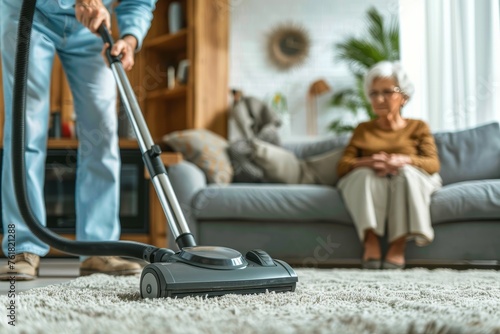 A person vacuuming a shaggy carpet, senior woman seated on the couch in the background, daily life captured