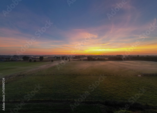 This wide-angle photograph captures the tranquil beauty of a sunrise spreading its warm glow over a lush pasture. The horizon is adorned with an array of colors, ranging from deep purples to fiery