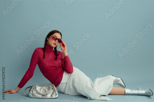 Fashionable confident woman wearing pink wraparound sunglasses, turtleneck, white denim maxi skirt, silver ankle boots, posing on blue background. Studio fashion portrait. Copy, empty space for text