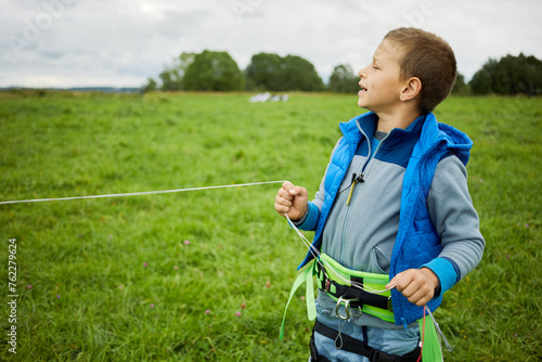 Little boy equipped for skysurfing stands holding flying lines in hand on green meadow.