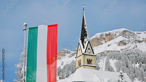 Church with Italian flag in Arabba, Italy (province Belluno). Dolomites mountains in the background with a blue sky and lots of snow, after heavy snowfall. 
