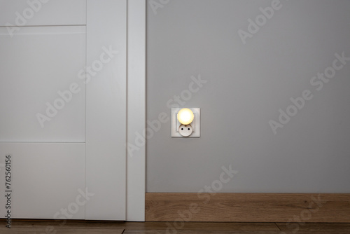 Obok drzwi znajduje się gniazdko z zasłonką emitującą światło, co symbolizuje połączenie konceptu elektryczności z ekonomicznymi zagadnieniami, zwłaszcza w kontekście wysokich rachunków za energię ele