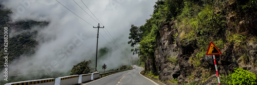 Winding mountain road with falling rocks warning sign amidst lush greenery and mist, ideal for adventure and travel-themed projects with ample copy space on the left