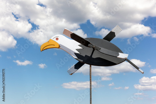 Seagull-shaped wooden weathervane on a thin rod in blue sky with white clouds background.