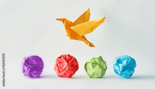 A photo of an origami bird flying over four differently colored crumpled paper balls on a white background, symbolizing creativity and imagination in business idea.