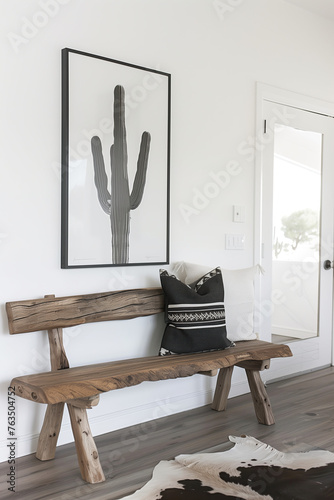 A rustic wooden bench with black and white cushion against the wall in modern farmhouse style home entryway, above it is hanging cactus artwork frame print on the wall, cowhide rug, white walls