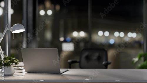 A laptop computer on a desk in a modern private office at night, illuminated by a table lamp.