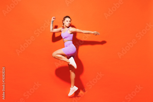 Full body side profile view young fitness trainer instructor woman sportsman wears top shorts purple clothes in home gym jump high isolated on plain orange background. Workout sport fit abs concept.