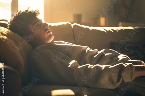 young man in sweats lying on couch, backlit with daylight