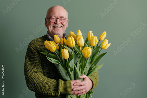 Joyful bald gentleman with a beaming smile holds a generous bouquet of yellow tulips, exuding warmth and happiness against a soothing green backdrop. Romantic senior man