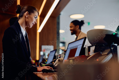 Smiling hotel manager working on computer at reception desk.