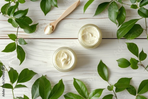 Natural deodorant creams in open jars with a wooden spatula and fresh green leaves on a white wooden background.