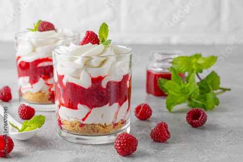 Raspberry trifle with whipped cream, berry sauce, cookie crumbs and mint leaves in a glass on a gray concrete background. Summer diet dessert.