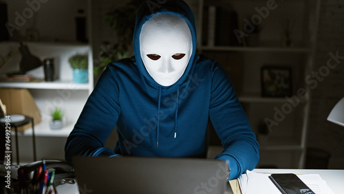A incognito man in a hoodie works intently on a laptop at night in a home office.