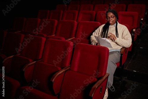 Minimal portrait of Black adult woman reading script in audience at theater in low light copy space