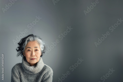 femme retraitée asiatique, origine japonaise, avec les cheveux gris, blancs et noirs, poivre et sel, avec un pull gris à col roulé face objectif, maquillée sur fond gris avec espace négatif copy space