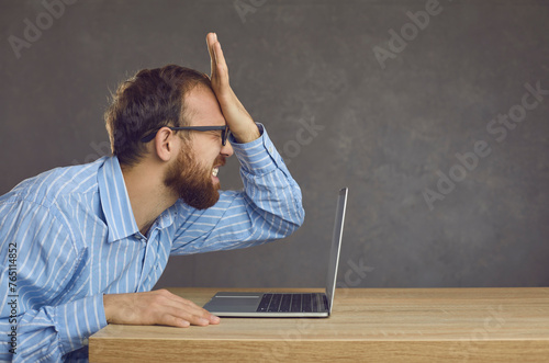 Argh, don't remember it. Side profile view angry ashamed guy slaps face sitting at office desk with laptop. Businessman makes stupid mistake online, forgets password, fails to fix computer malfunction