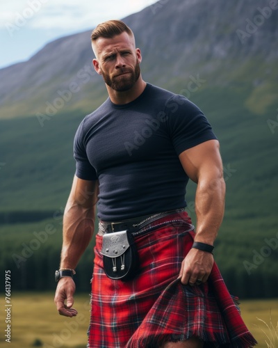 handsome muscular Scottish man dressed in a T-shirt and red plaid kilt kilt stands against the background of mountains