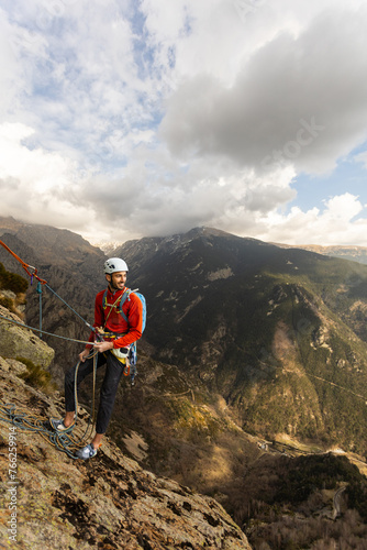 A man in a red jacket is standing on a mountain with a backpack. He is wearing a helmet and he is enjoying the view