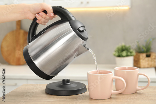 Woman pouring hot water from electric kettle into cup in kitchen, closeup