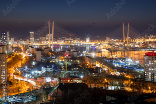 Beautiful night city landscape. Top view of streets and buildings. In the distance is a large cable-stayed Golden Bridge over the Golden Horn Bay. Vladivostok city, Primorsky Krai, Russian Far East.