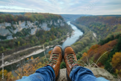 A tranquil scene unfolds as a hiker sits on the edge of a cliff, feet in sturdy hiking shoes against the backdrop of a winding river and the autumnal colors of the forest.
