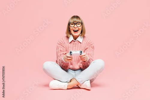 Full body elderly woman 50s years old wear sweater shirt casual clothes glasses sits hold in hand play pc game with joystick console isolated on plain pastel light pink background. Lifestyle concept.