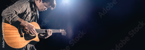 A male guitarist plays an acoustic guitar on a dark stage background.