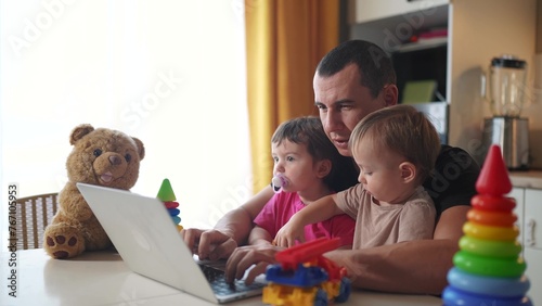 father working from home a remotely with two baby in fun his arms. pandemic remote work business concept. father tries to work at home in kitchen, baby children interfere sitting on their hands