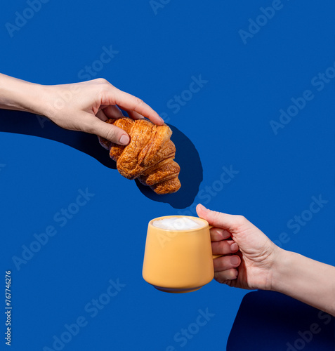 Women's hands hold fresh croissant and cup of coffee on a blue background.