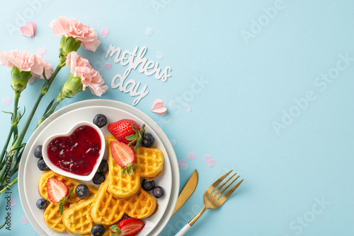 Mother's Day breakfast table: top view heart waffles, strawberries, blueberries, sweet syrup, cutlery, carnations, note "mothers day," on a light blue surface, area for message