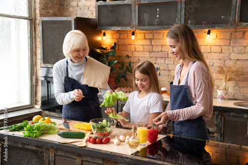 Horizontal shot of smiling family cooking together in home kitchen. Vegetables and salad on desk. Daughter, mother and grandmother preparing fresh organic vegetables for salad