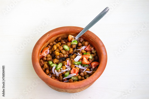 Boiled chickpeas and spoon in an earthen pot on white wooden textured background. Chopped tomatoes, onion, and green chilies mixed with Bengal gram. Healthy weight loss meal. 