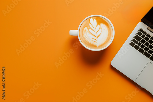 tasse à café blanche remplie d'un art caffe latte à la mousse blanche, à côté d'un ordinateur portable ouvert, laptop et café sur un fond orange espace négatif copy space