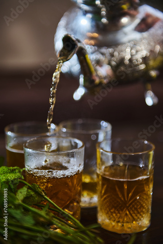 Pouring delicious Moroccan mint tea from a silver teapot into drinking glasses. Bunch of fresh mint in the foreground