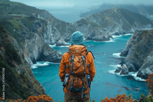 An individual hiker in an orange jacket stands taking in the view over a dramatic coastline with steep cliffs and the open sea
