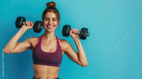 Athletic smiling woman in top holding dumbbells looking at camera, pumping biceps at gym. Indoor studio shoot isolated on blue background