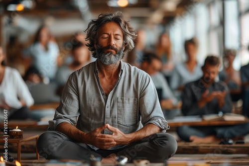 A man with a beard and moustache is sitting in a lotus position with his eyes closed, participating in a meditation event in front of a group