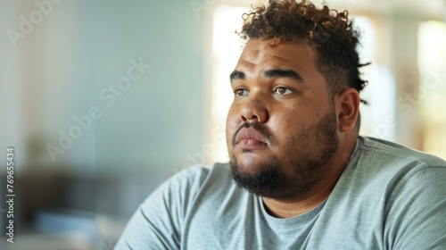 A man with a beard and curly hair wearing a gray t-shirt sitting in a room with a blurred background looking contemplative.