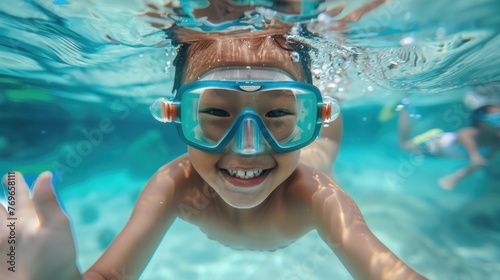 A young child with a big smile wearing blue goggles swimming underwater in a clear blue pool.