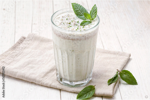 A glass of Turkish traditional drink, ayran, kefir, or buttermilk made from yogurt, garnished with mint leaves, is placed on a beige napkin on a white wooden table.