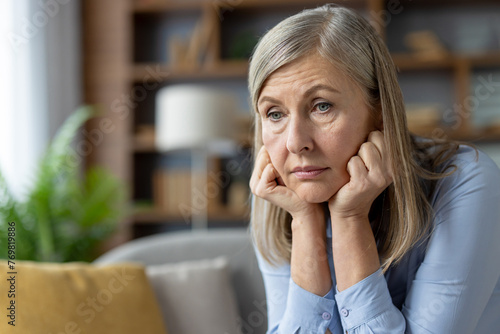 A middle-aged woman appears worried, sitting alone on a couch in a comfortable home setting, her hands on her head.