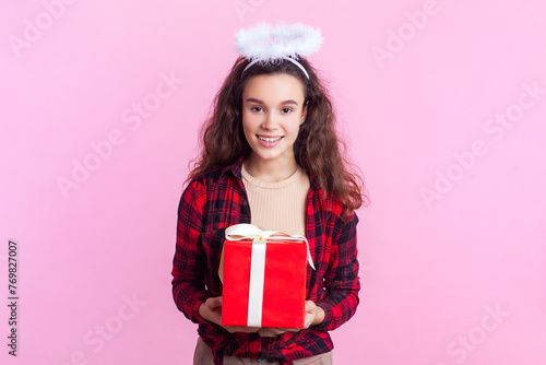 Portrait of pretty pleased teenage girl with wavy hair in red checkered shirt and nimb over head standing giving present box charity. Indoor studio shot isolated on pink background.