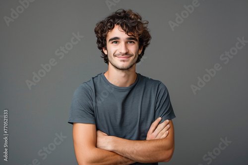 Photo of a happy young man with curly hair in a gray shirt standing on a gray background with his arms crossed and looking at the camera smiling