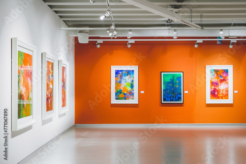 An airy white art gallery with an orange wall, creating a warm, inviting atmosphere. White frames contain vibrant, abstract paintings, their bright colors standing out against the orange. 