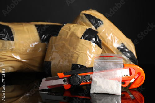 Smuggling, drug trafficking. Packages with narcotics and utility knife on black mirror surface, closeup