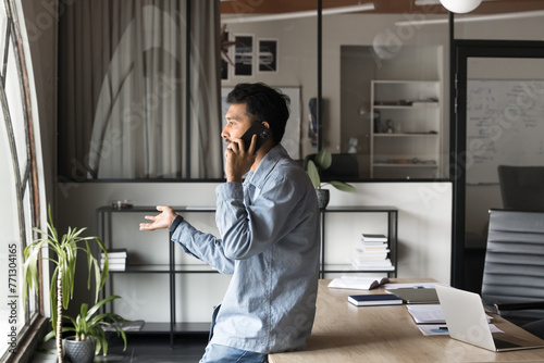 Serious young Indian business professional man in casual speaking on cellphone in office, leaning at workplace table looking at window away, talking to customer, moving hands. Side view